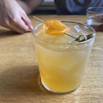 Calico - A yellow cocktail with ice cubes and a fruit garnish on a wooden table, with two people in the background, one holding a glass of water. - Cocktail Bars Near Me - Washington, DC, District of Columbia