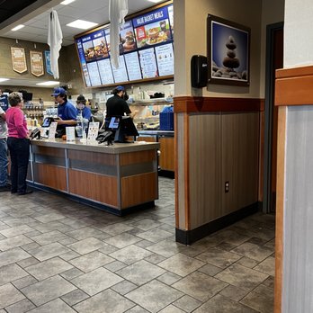 Culver's - The image shows the interior of a fast-food restaurant with digital menu boards displaying meal options. Customers are standing at the counter placing orders, while staff members are preparing food behind the counter. The decor includes wood paneling and tiled flooring, with umbrellas hanging from the ceiling. A framed picture of stacked stones is mounted on the wall, and a speaker is attached nearby. - Fast Food Near Me - Mesa, Arizona