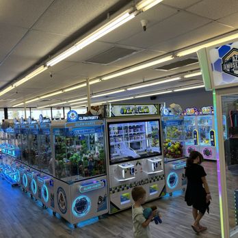Claw Party Arcade - The image shows an indoor amusement arcade with multiple claw machines filled with plush toys. Several children are engaged with the games, and an adult is seen walking nearby. The arcade has bright overhead lighting, and the machines are decorated with colorful signage and LED lights. The setting appears to be a family-friendly entertainment center with a variety of arcade games. - Arcades Near Me - Glendale, Arizona