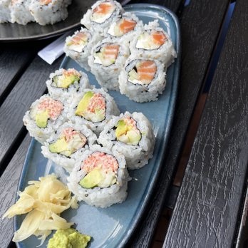 The Roost - California and salmon and cream cheese roll - A rectangular blue ceramic plate holds two types of sushi rolls: one with salmon, cream cheese, and cucumber, and another with crab, avocado, and possibly shrimp. The rolls are neatly arranged on a dark wooden table, with a small portion of pickled ginger and wasabi placed at the corner of the plate. In the background, a black plate with additional sushi rolls is partially visible, suggesting a shared dining experience at an outdoor or casual restaurant setting. - Coffee & Tea Near Me - Washington, DC, District of Columbia