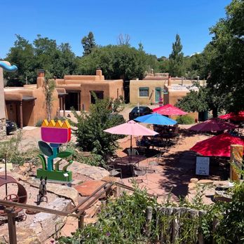 Cafe Canyon - Patio view from sidewalk - Outdoor patio with multiple tables and chairs shaded by pink, blue, and red umbrellas, situated among adobe-style buildings with a desert landscape. The area is surrounded by greenery, including trees and bushes, with a clear blue sky overhead. The scene suggests a relaxed dining or cafe setting in a southwestern-style environment. - Coffee & Tea Near Me - Santa Fe, New Mexico