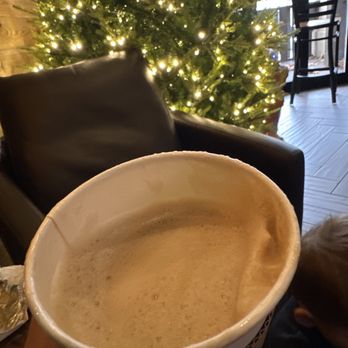 Southdown Coffee - Patchogue - chia latte - A hand holds a cup of coffee with foam on top, positioned in the foreground of the image. Behind the cup, a decorated Christmas tree with string lights is visible, along with a black leather chair and a window showing an outdoor scene. The setting appears to be a cafe with wooden flooring and furniture, creating a warm, festive atmosphere. - Coffee Roasteries Near Me - Patchogue, New York