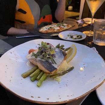The Front Yard - A restaurant setting with a plated seafood dish consisting of a piece of fish topped with microgreens, accompanied by cooked asparagus and a lemon wedge. The dish is presented on a white plate with a gold rim, placed on a dark table. In the background, there are additional plates, a cocktail glass, and a glass of water, with a person partially visible wearing a black shirt with a colorful abstract design. - Mediterranean Near Me - Ogunquit, Maine