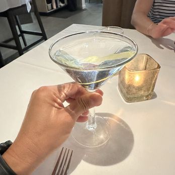 Prezza - Vodka martini - A hand holding a martini glass with a clear cocktail, possibly a vodka martini, on a restaurant table with a fork and a candle holder. - Italian Near Me - Boston, Massachusetts