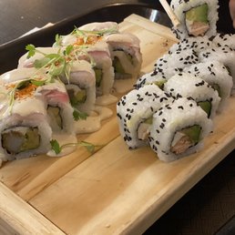 California roll and shrimp tempura roll - Two types of sushi rolls are displayed on a wooden serving board, one topped with sauce and microgreens, the other with black sesame seeds, with chopsticks holding a piece.