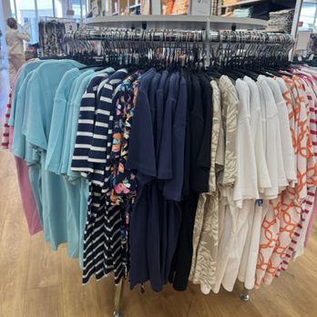 Chico's Off The Rack - A round clothing rack holds an assortment of polo shirts and t-shirts in different colors and patterns, located inside a retail store with shelves of folded clothes in the background. - Women's Clothing Near Me - Estero, Florida