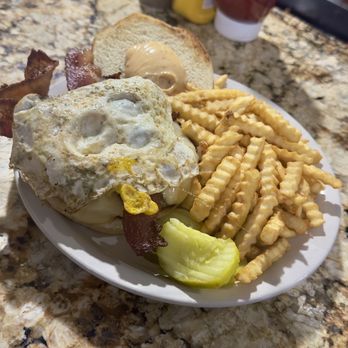 Outpost - The Jimmy - A breakfast dish featuring a fried egg on bacon, waffle fries, pickles, a bread slice with mayonnaise, and condiments in the background. - American Near Me - Anoka, Minnesota