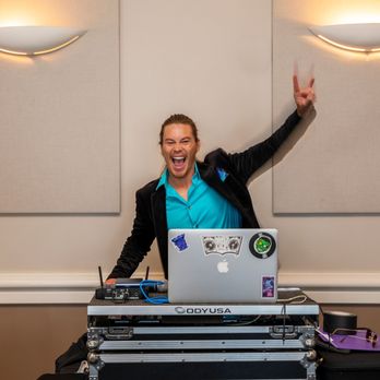 VOX DJs - Caleb Riley at our wedding! - A man with long hair, wearing a bright blue shirt and black jacket, is standing behind DJ equipment with a laptop, smiling and making a peace sign. - Photo Booth Rentals Near Me - Costa Mesa, California