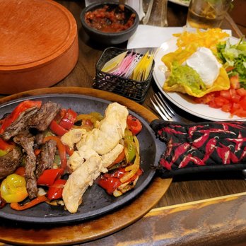 El Bruno's - Fajitas - The image shows a meal set on a wooden table, featuring a sizzling plate of fajitas with beef strips, red and green bell peppers, and fried strips of chicken. A side salad with chopped lettuce, tomatoes, shredded cheese, and sour cream is on a white plate. Two glasses of water and a glass with a beverage are also visible. Condiments and utensils are present, with a black oven mitt placed next to the fajitas. - Mexican Near Me - Albuquerque, New Mexico