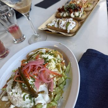 Hash Kitchen Arcadia - Kristen is the best bartender  Will be back because of her - A white oval plate contains chilaquiles topped with pickled red onions, crumbled cheese, and cilantro, served with fried tortilla chips. In the background, there are tacos with various toppings, including sour cream, salsa, and greens, on a rectangular tray. The table has drinks, including a martini glass with a creamy beverage, and other glasses with pink and red drinks. Black napkins are also on the table, indicating a casual dining setting. - Breakfast & Brunch Near Me - Phoenix, Arizona