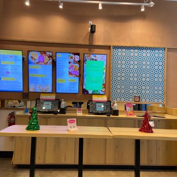 Taim - The image shows a checkout counter with four digital screens displaying menus, set against a wooden wall with a decorative blue patterned panel. Small Christmas trees and holiday decorations are placed on the counter, indicating a festive season. The area includes a cash register, hand sanitizer, and signage, with warm lighting overhead and a clean, organized appearance. - Mediterranean Near Me - Brooklyn, New York