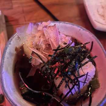 Kinda Izakaya - Agedashi tofu - A bowl of agedashi tofu, a Japanese dish, is shown with toppings including bonito flakes and shredded nori. The tofu is in a dark soy-based sauce, and the bowl is placed on a wooden table. The setting appears to be a Japanese izakaya restaurant, with a partial view of another dish in the background. - Tapas/Small Plates Near Me - Berkeley, California