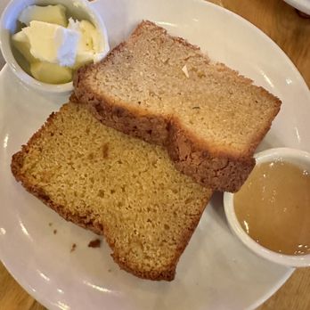 M'tucci's Italian - Extra virgin olive oil cake with butter and Bee Chama honey - Two slices of cornbread are placed on a white round plate. One slice appears slightly darker and more textured, while the other is lighter and more uniform. Two small bowls contain butter with honey and butter with a light-colored syrup, positioned near the cornbread. The plate is on a wooden table, with part of another dish visible in the background. - Italian Near Me - Albuquerque, New Mexico
