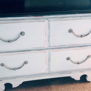 Juxtaposed - A white wooden dresser with four drawers, each featuring metal handles, is positioned on a carpeted floor. The dresser has a distressed finish with visible wear and tear, and a dark-colored top surface. The design includes curved cutouts at the bottom and a slightly aged appearance. - Home Decor Near Me - Albuquerque, New Mexico
