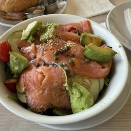 Smoked Salmon Salad - A white ceramic bowl holds a salad with slices of smoked salmon, avocado, cucumber, cherry tomato, and capers. The salad is placed on a light wooden table, with a plate of a sandwich and utensils visible in the background. The ingredients are fresh and arranged in a casual presentation.