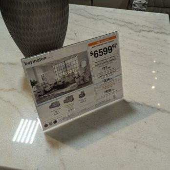 Ashley Outlet - A price tag for furniture is displayed on a marble table, with a large vase behind it and a person in sneakers visible in the background. - Furniture Stores Near Me - Jackson, Michigan