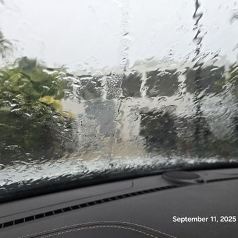 Jonathan Window Tint - A view through a car windshield during rain, with water droplets and streaks obscuring the scenery outside, including trees and cloudy sky. - Home Window Tinting Near Me - Boca Raton, Florida