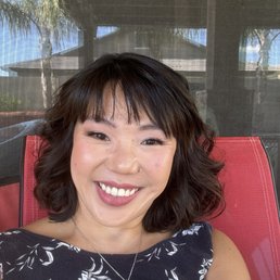 A woman with dark, wavy hair and a floral-patterned top is smiling at the camera while sitting outdoors on a red chair. The background shows a screened porch with a roof, trees, and a building, indicating an outdoor setting. She is wearing a necklace with a pendant and appears relaxed and happy.