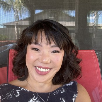 Le Boudoir Studio - A woman with dark, wavy hair and a floral-patterned top is smiling at the camera while sitting outdoors on a red chair. The background shows a screened porch with a roof, trees, and a building, indicating an outdoor setting. She is wearing a necklace with a pendant and appears relaxed and happy. - Boudoir Photography Near Me