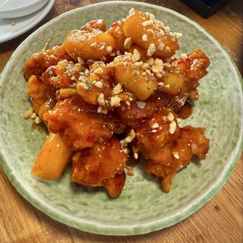 MORU - Fried chicken (Korean style ~) - A dish featuring Korean-style fried chicken pieces coated in a glossy, orange-colored sauce, garnished with chopped peanuts. The chicken is served on a green ceramic plate, with some potato chunks visible underneath. The setting appears to be a restaurant, with a wooden table and some empty dishes nearby. - Japanese Near Me - Fort Lee, New Jersey