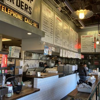 Willards Real Pit BBQ - The image shows the interior of a barbecue restaurant with a counter where customers place orders. Overhead, large menu boards display various food options, including BBQ sandwiches and platters. Customers are seen standing and waiting, while staff members work behind the counter. The decor includes industrial-style lighting and signage, with a soda fountain visible on the left side. The setting appears casual and busy, with a focus on takeout and dine-in service. - Barbeque Near Me - Reston, Virginia