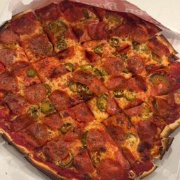 A large pizza with a thin crust, topped with pepperoni slices, jalapeño peppers, and melted cheese, is placed in an open cardboard pizza box on a white surface. The pizza appears freshly baked with slightly crispy edges and a variety of toppings evenly distributed across the surface.