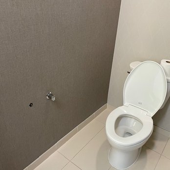 SpringHill Suites by Marriott Durango - broken toilet paper holder - A bathroom features a white ceramic toilet with the lid open, situated against a wall with a gray textured wall on one side and a plain light-colored wall on the other. A roll of toilet paper is placed on top of the tank, and a small pipe or fixture is visible on the gray wall. The floor is tiled with light-colored tiles. - Hotels Near Me - Durango, Colorado