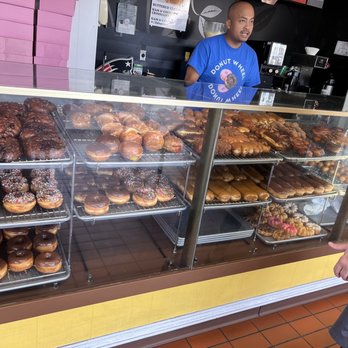Donut Wheel - a man behind the counter of a donut shop - A glass display case contains multiple shelves of donuts, including glazed, sprinkled, chocolate, and filled varieties, at Donut Wheel in Chico, California. A staff member wearing a blue Donut Wheel shirt is behind the counter, and a customer is pointing at the donuts. The shop has a vintage-style decor with a black and red color scheme and a tiled floor. - Donuts Near Me - Chico, California