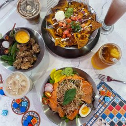 The image shows a white marble table with multiple dishes and drinks. There are nachos topped with cheese, tomatoes, guacamole, and sour cream, a plate of fried chicken pieces with a yellow dipping sauce, and a salad with smoked salmon, radishes, cherry tomatoes, and a boiled egg. Beverages include iced coffee, iced tea, and a pink milkshake. A board game with colorful tiles and pieces is partially visible on the right side of the table. The setting appears casual and inviting, suitable for a meal and game session.
