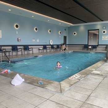 SpringHill Suites by Marriott Madison - The image shows an indoor hotel pool with a girl swimming in the water. The pool area has a tiled deck with a towel and some toys on the edge. Along the wall, there are multiple chairs and a person sitting on the floor near a door. The wall is painted light blue with circular wall lights, and a clock is visible on the wall. The ceiling is dark with a wood-like finish, and there are signs and a warning notice near the pool. - Hotels Near Me - Madison, Wisconsin