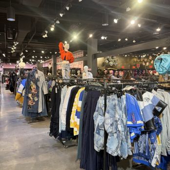Akira - The image shows the interior of a retail clothing store with multiple racks filled with casual and sportswear clothing, including jackets, shirts, and pants. Mannequins are positioned on elevated platforms, dressed in outfits, with one mannequin wearing an orange jacket and light-colored pants. The store has a modern industrial ceiling with numerous spotlights illuminating the space. Shoppers are visible in the background browsing and shopping, and the store appears spacious with a variety of clothing options. - Women's Clothing Near Me - Paramus, New Jersey