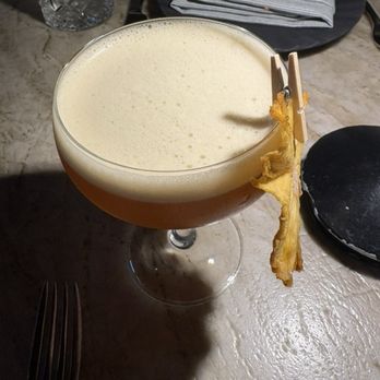 Rumis Kitchen - Gifts of the Beloved - Cocktail. Really nice. - A foam-topped cocktail is presented in a coupe glass, garnished with a strip of bacon clipped to the rim, on a marble table. - Persian/Iranian Near Me - Houston, Texas
