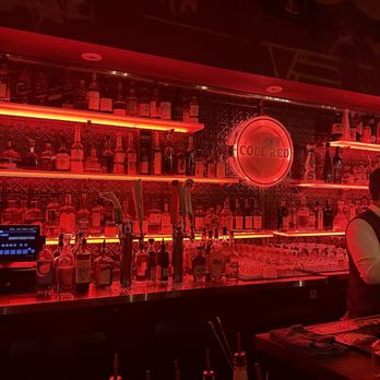 CODE RED - The image shows a bar with illuminated shelves filled with bottles of liquor, highlighted by red lighting. A neon sign reading 'CODE RED' is centered on the wall behind the bar. A bartender is visible on the right side, preparing drinks, with bar tools and glasses on the counter. The overall ambiance is dark and moody, emphasizing the red lighting and the bar's decor. - Cocktail Bars Near Me - Washington, DC, District of Columbia