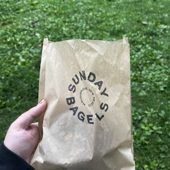 Sunday Bagels - A hand holds a crumpled paper bag with "Sunday Sweets Bagel" printed on it, against a background of green grass. - Bagels Near Me - Albuquerque, New Mexico