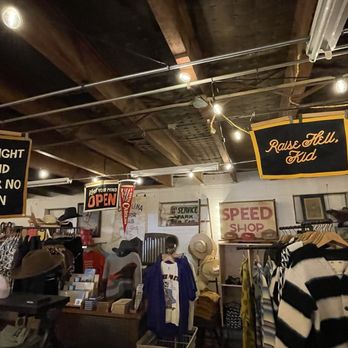 Unlikely Professionals - Inside - The image shows the interior of a gift shop with a wooden ceiling and various hanging signs, including motivational quotes. Shelves and racks hold hats, clothing, and accessories, with some items on display tables. The shop has a casual, eclectic atmosphere with diverse merchandise and decorative elements. - Men's Clothing Near Me - Raleigh, North Carolina