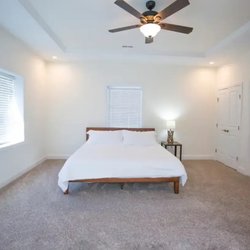 Premiere Renovations And Construction - A spacious bedroom features a wooden-framed bed with crisp white linens, a small nightstand with a lit lamp, tray ceiling, ce...