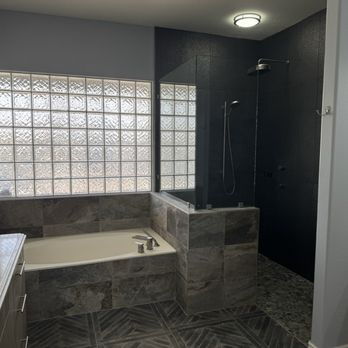 Marshall Renovations - Relaxing spa space - Bathroom includes a built-in bathtub with a marble tile surround, positioned under a large glass block window. Adjacent is a walk-in shower with black textured tiles, a rainfall showerhead, and a handheld shower. The floor has dark, patterned tiles, and the ceiling has a modern light fixture. The space combines natural light and contemporary design elements. - General Contractors Near Me