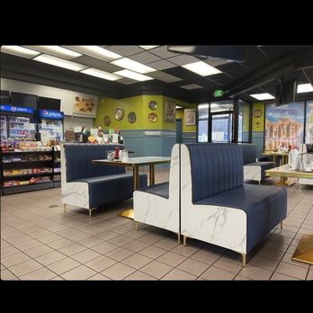 Sultan - A view of the dining area, and the checkout counter - A restaurant interior with booth seating arranged around tables, featuring navy blue cushions and white marble-like sides. The floor is tiled, and the ceiling has rectangular light panels. In the background, there are Pepsi refrigerators stocked with snacks and drinks, and the walls are decorated with plates and framed artwork. Large windows and an exit door are visible, providing natural light and views outside. - Falafel Near Me - Breezewood, Pennsylvania