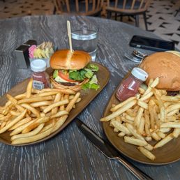 STRAUSS GRASS-FED CHEESEBURGER w/ fries - Two cheeseburgers with fries are served on wooden plates, accompanied by ketchup and water glasses, in a restaurant setting.