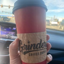A person is holding a large coffee cup from Grunders Coffee House inside a car. The cup has a brown sleeve with the Grunders logo and is topped with a black lid. The background shows the car's dashboard and a view of traffic and blue sky through the windshield.