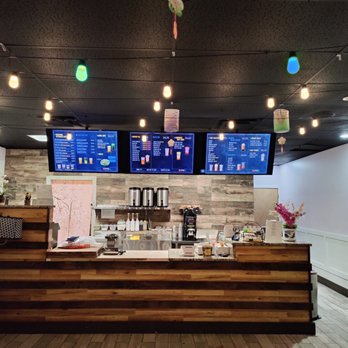 KUTEA - a coffee shop counter with a variety of menus - The image shows the interior of a Vietnamese-style boba shop with a wooden counter and three digital menu screens displaying drink options. The ceiling is decorated with string lights and paper lanterns. The background includes a wall with wood paneling, a small plant, and a pink decorative element. The shop has a modern yet cozy atmosphere suitable for enjoying bubble tea. - Desserts Near Me - Denver, Colorado