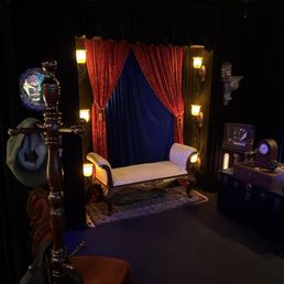 The image shows a small, vintage-style stage with red velvet curtains tied back, revealing a white upholstered bench with ornate wooden legs. The stage is decorated with warm, yellow wall-mounted lights and a dark backdrop, creating an intimate atmosphere. To the right, there is a dark cabinet with decorative items, and a small television or monitor. The room has dark walls and ceiling, with a few vintage-style light fixtures, suggesting a cozy, theatrical environment.
