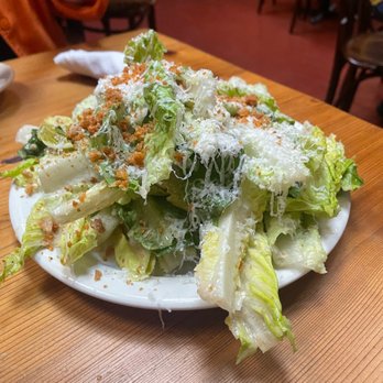 The Fly - A Caesar salad with chopped romaine lettuce, grated cheese, and croutons is presented on a white plate. The salad appears fresh with a generous topping of cheese and crunchy croutons, placed on a wooden table in a restaurant setting. The background shows chairs and tables, indicating a dining environment. - Wine Bars Near Me - Brooklyn, New York