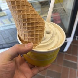 Vanilla soft serve ice cream in a yellow cup, topped with a waffle cone piece, held by a hand inside a store.