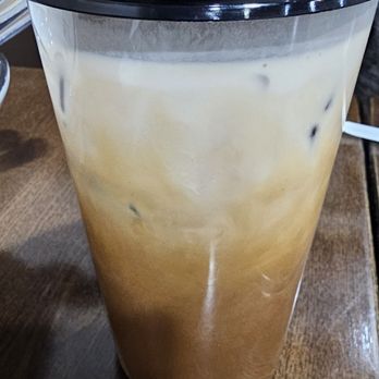 Pho Table - Bath Beach - A tall, transparent plastic cup filled with iced coffee mixed with milk and ice cubes is shown on a wooden table. The drink has a layered appearance with darker coffee at the bottom and lighter milk at the top. The cup is covered with a black plastic lid. The setting appears to be a café or casual dining area. - Vietnamese Near Me - Brooklyn, New York