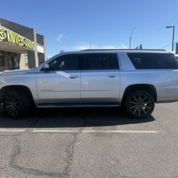 Malone's Quality Tire - A silver SUV is parked in a parking lot in front of a store with a sign indicating it sells guns, knives, and ammunition. The vehicle has dark tinted windows and black wheels. The store's exterior is made of stone and has large yellow and black signage. The sky is clear and blue, with some power lines and traffic lights visible in the background. - Tires Near Me - Apache Junction, Arizona
