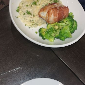 110 Grill - A white bowl holds mashed potatoes topped with chopped herbs, accompanied by steamed broccoli florets and a piece of meat wrapped in bacon. The dish is presented on a dark table, with utensils visible nearby. - American Near Me - Hadley, Massachusetts