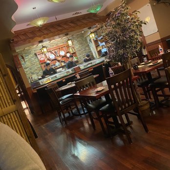 Nori - a restaurant with a wood table and chairs - The image shows the interior of a Japanese-inspired restaurant with a sushi bar where chefs are preparing dishes. The space includes wooden tables and chairs arranged for diners, and a large potted plant is positioned near the seating area. The ceiling is decorated with colorful umbrellas, and warm lighting creates a cozy atmosphere. The restaurant has a traditional aesthetic with a mix of modern and classic Asian design elements. - Japanese Near Me - Montclair, New Jersey