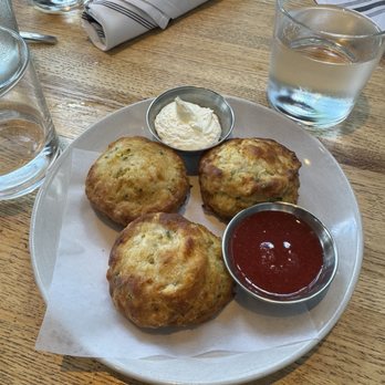 Showroom - Cheddar Chive biscuit with whipped butter and strawberry jam - A white plate holds three biscuits with a golden-brown exterior, served with whipped butter and strawberry jam in small metal containers. The plate is on a wooden table with glasses of water, a rolled napkin, and a striped napkin nearby, indicating a casual dining setting. - New American Near Me - Frederick, Maryland