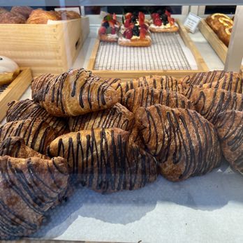 Paris Baguette - A pile of croissants with chocolate drizzle is arranged on a tray in a bakery display case, with other pastries visible in the background. - Bakeries Near Me - Chicago, Illinois